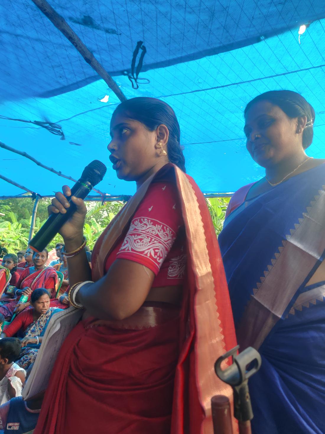 A woman speaks into a microphone under a blue canopy at the Mangrove Lab Exhibition.