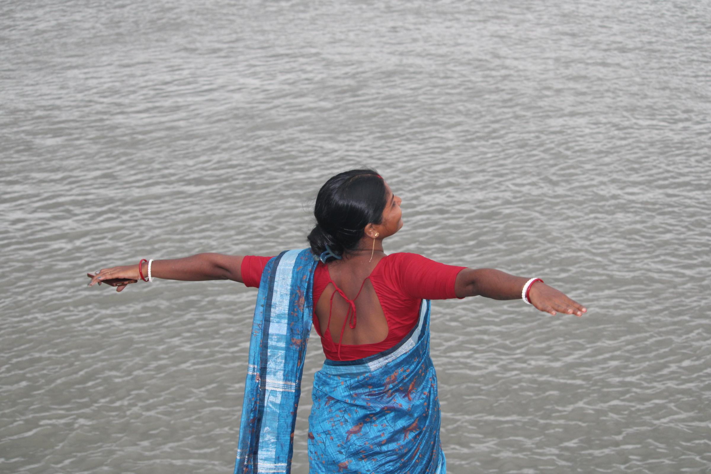 A woman in a blue sari stands at the river's edge on Bali Island, arms spread wide over the water.