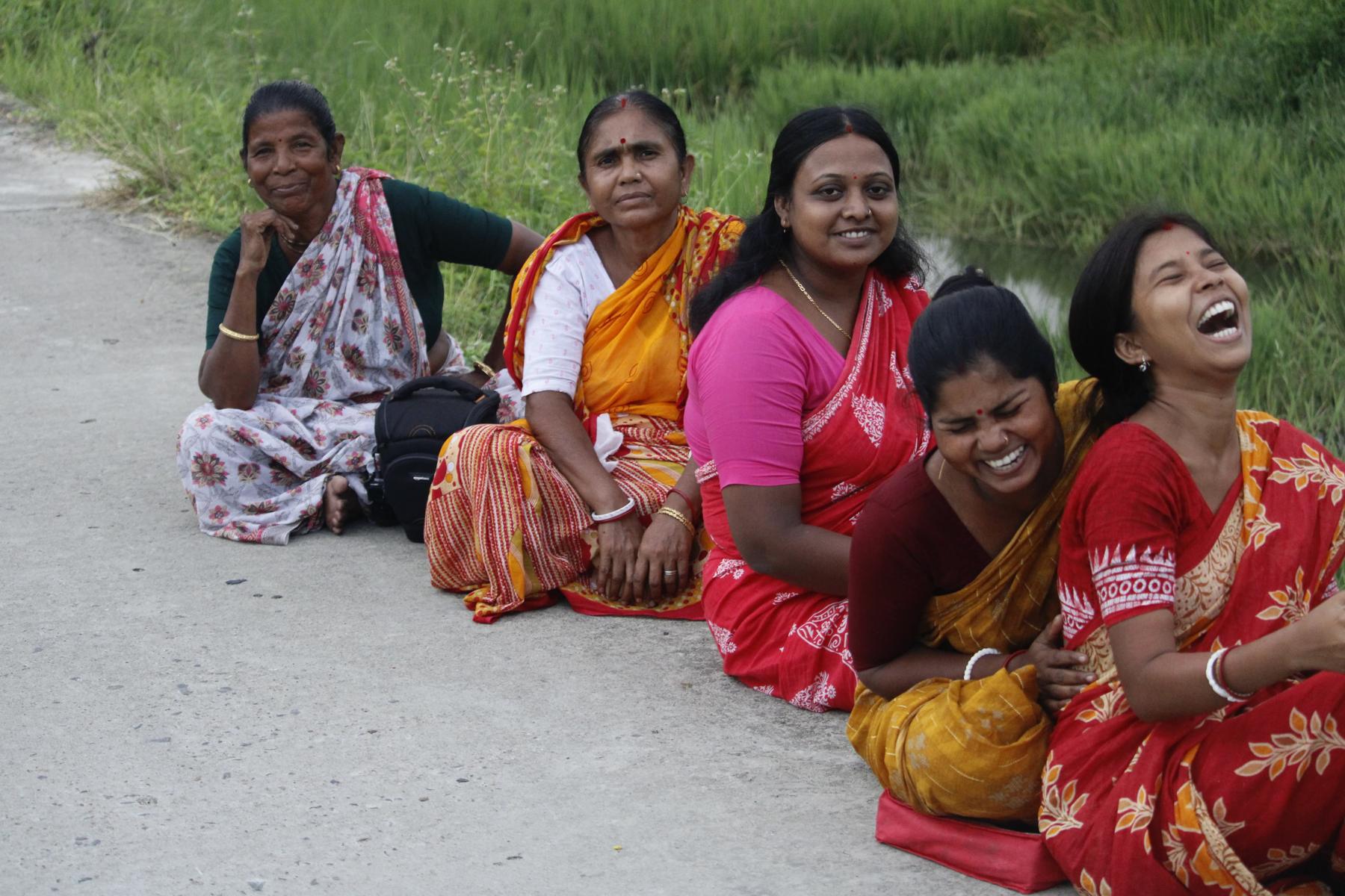 Women sitting together at the roadside beside a paddy field, laughing.